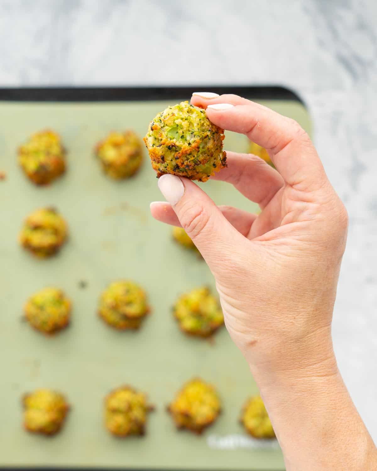 Cooked golden and crisp broccoli bites on a lined baking tray sitting on a bench with a hand holding one above them. 