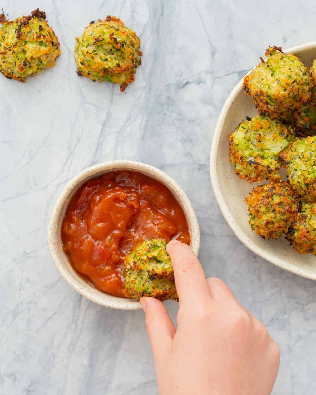 Baked broccoli bites in a serving dish with a hand dunking one into a ramekin of tomato salsa. 