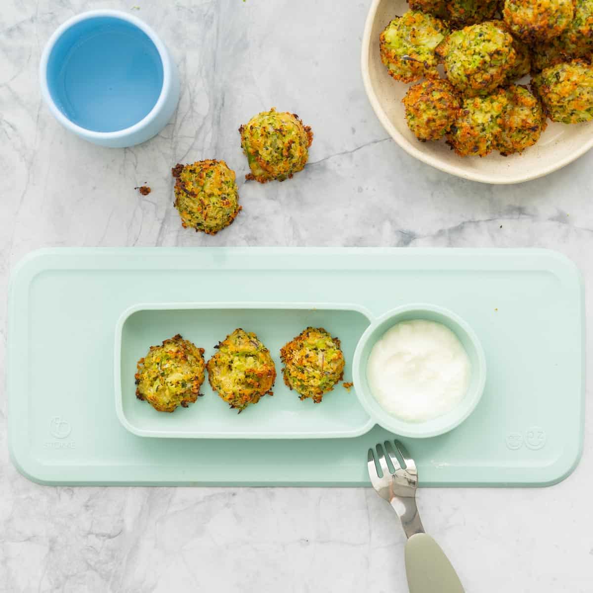 Baked broccoli bites line-up in a light green silicon toddler plate with a tablespoon of yoghurt and an additional serving bowl full of broccoli bites sitting on the bench