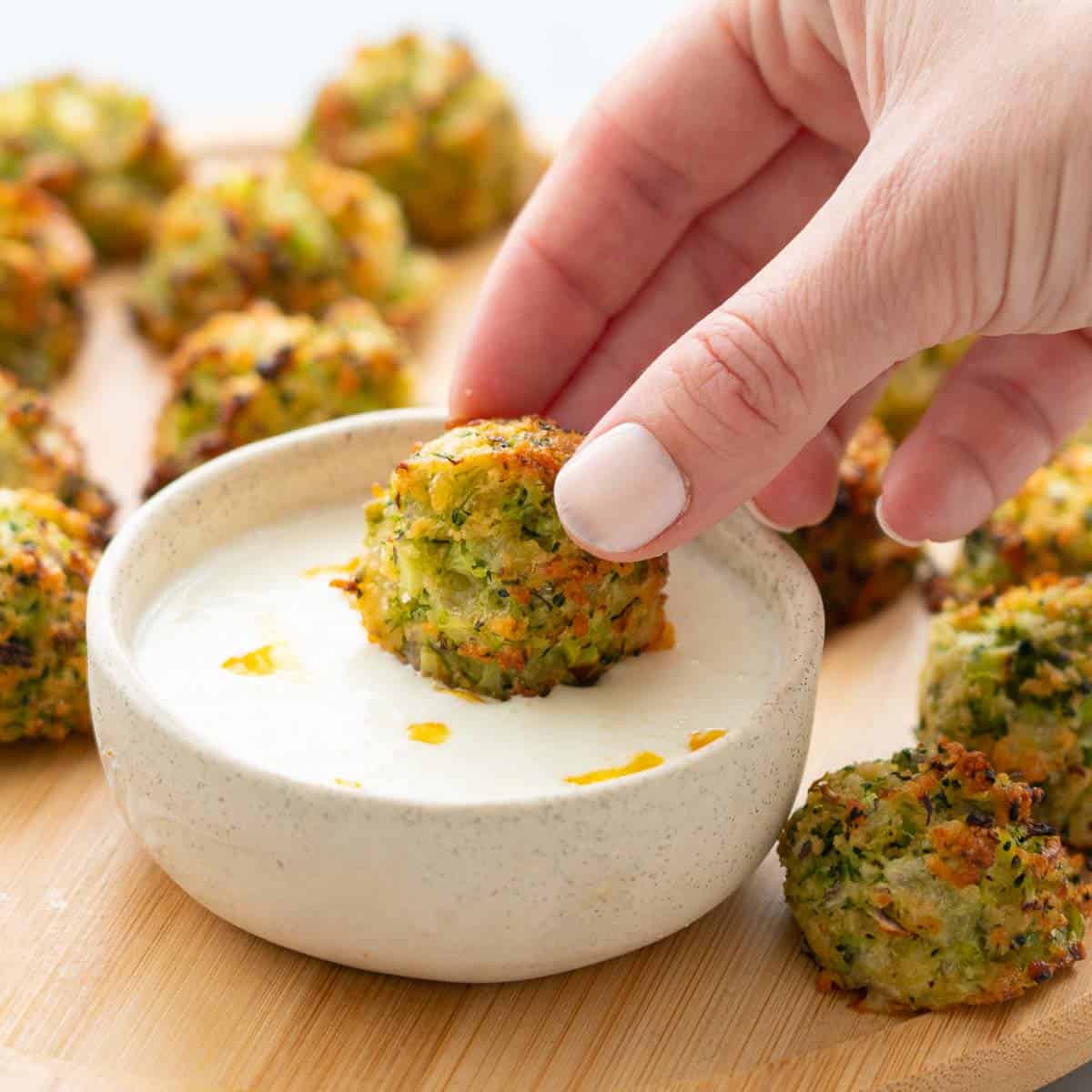 A hand dunking a broccoli bite into a ramekin of yoghurt and lemon dip which is sitting amongst more broccoli bites on a wooden serving board 
