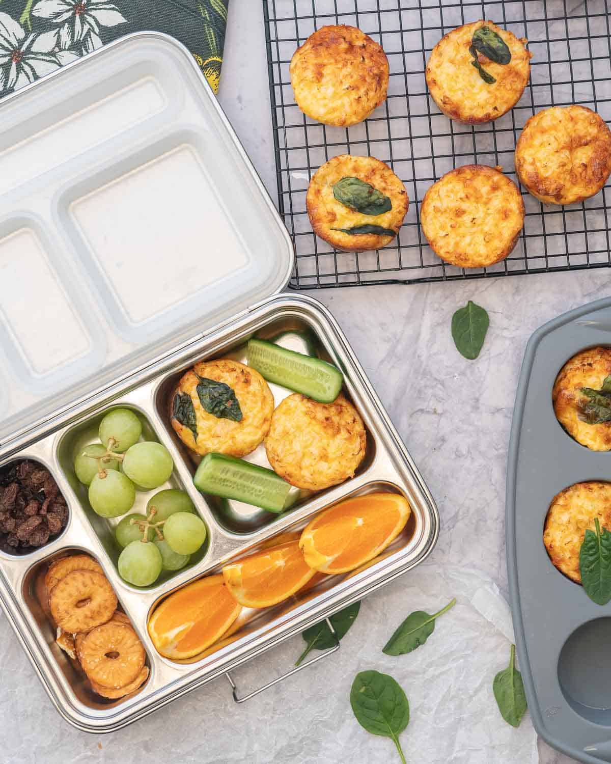 A tin lunch box filled with snack food and breakfast egg muffins sitting on a bench next to a tray and cooling rack of egg muffins 
