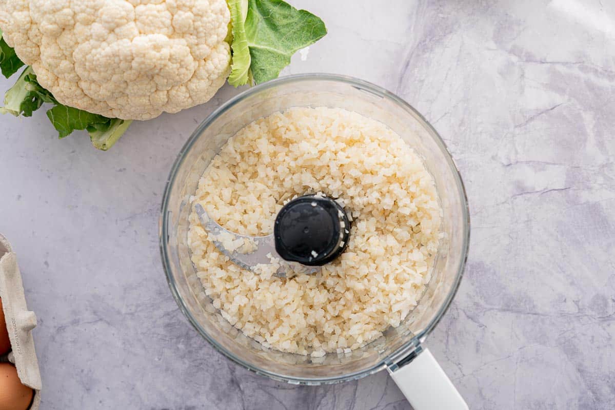 A food processor full of cauliflower rice sitting on the bench next to a large cauliflower 