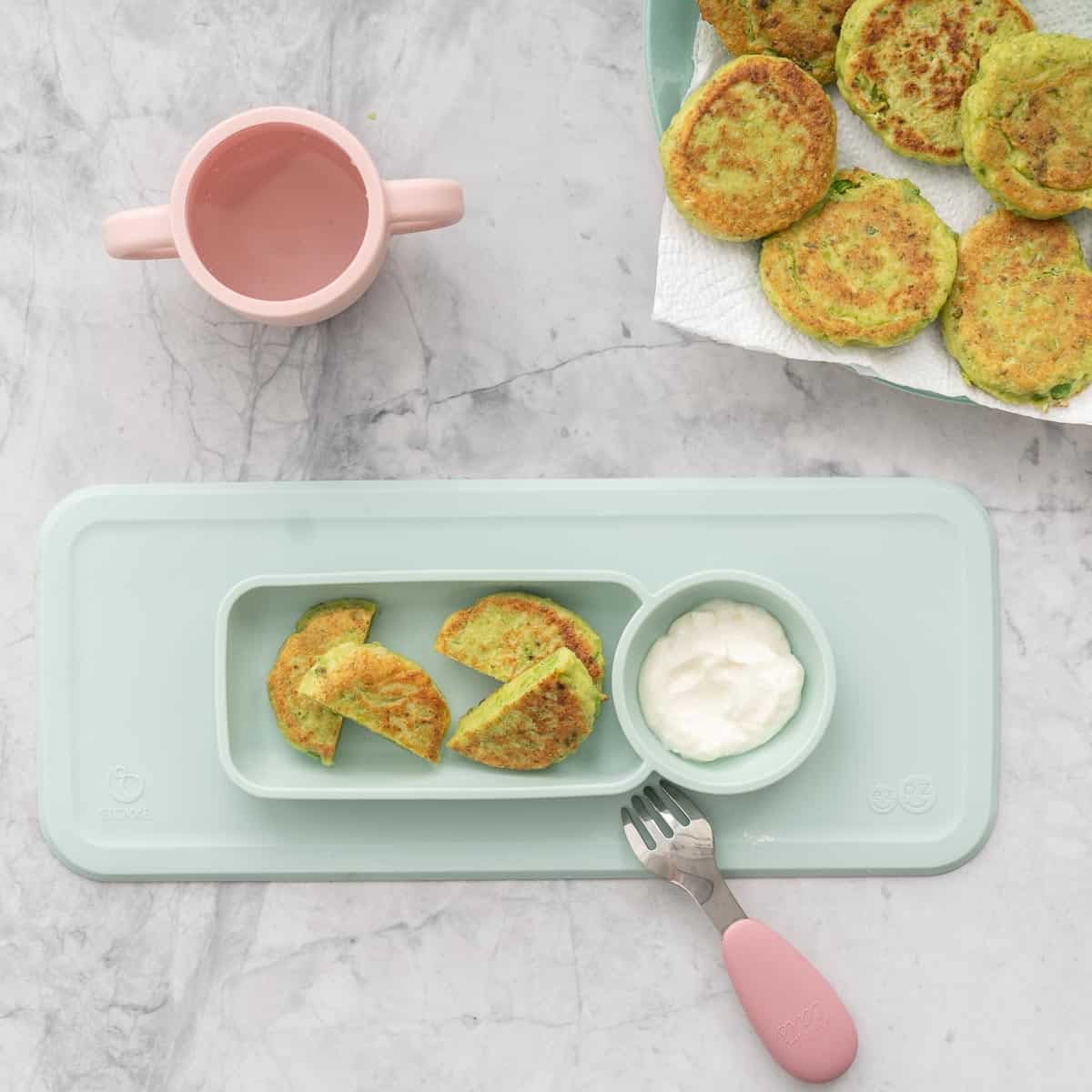 A silicon toddler plate with a serving of pea fritters and a dollop of greek yoghurt sitting on a bench next to a plate full of pea fritters in the background and a pink cup
