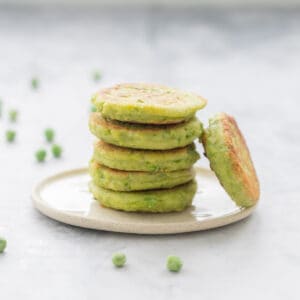 A tower of 6 small green fritters on a side plate with peas scattered around.