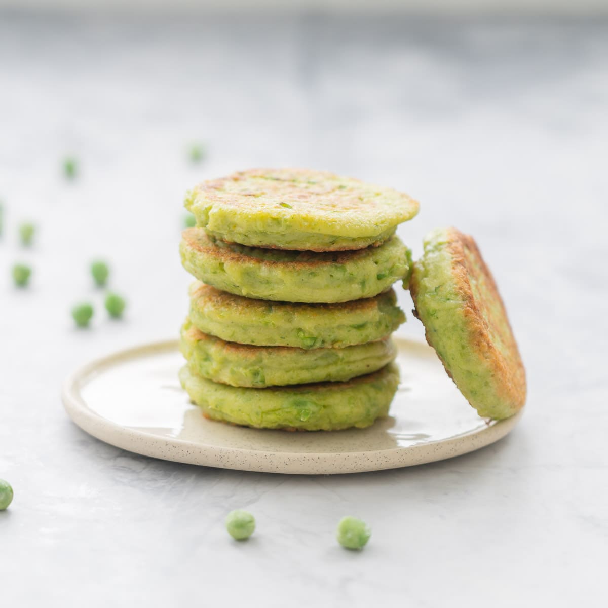 five golden pea fritters stacked on a plate with one leaning against them and a scattering of peas in the background on the bench