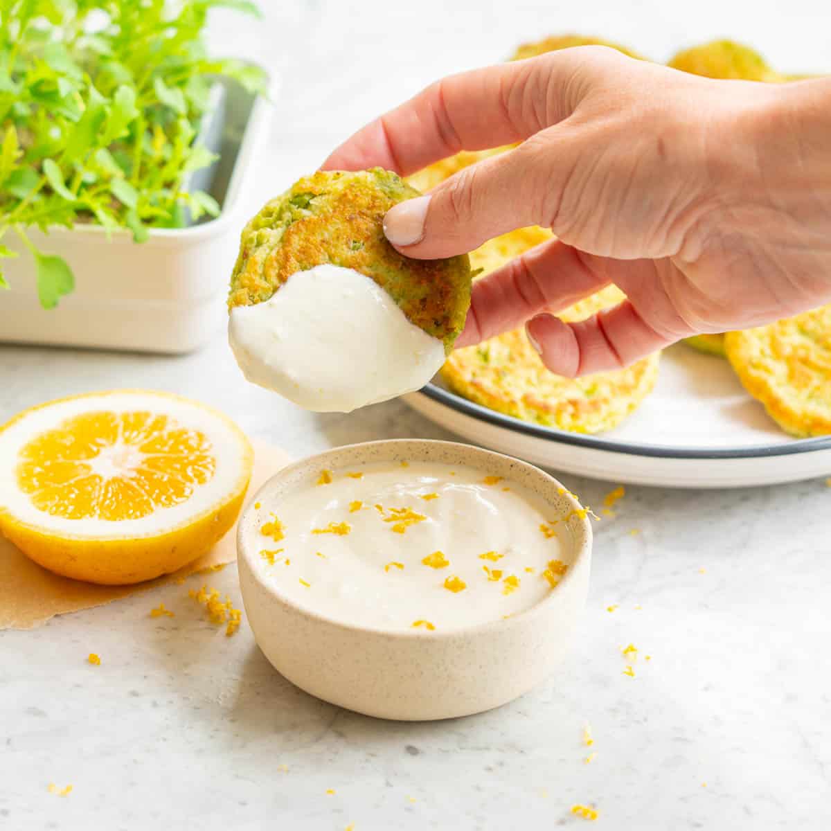 A hand holding a pea fritter which has been dipped into a yoghurt dressing which is sitting below next to a slice of lemon, a bowl full of green salad and a plate of pea fritters 