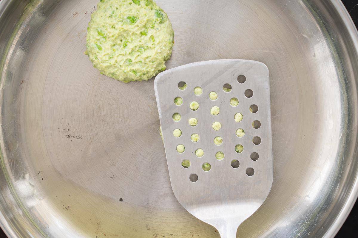 Two pea fritters cooking in an oiled frying pan with a spatular pressing down on one of them