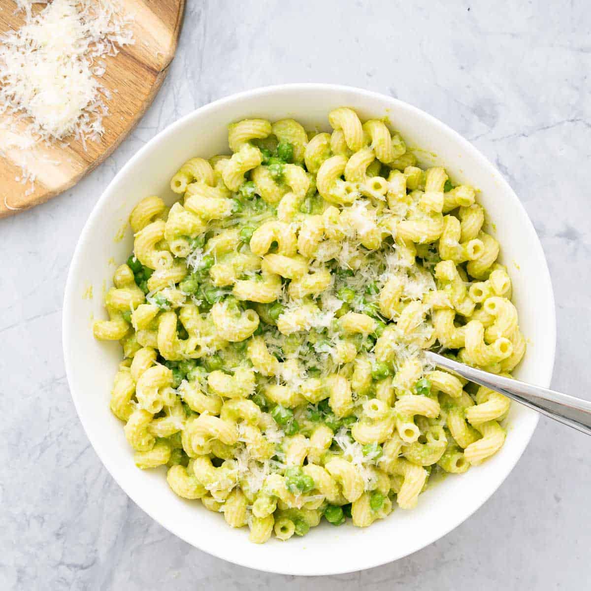 A large serving of Pea Pasta in a bowl sprinkled with parmesan cheese sitting on the bench with a spoon resting on the side. 