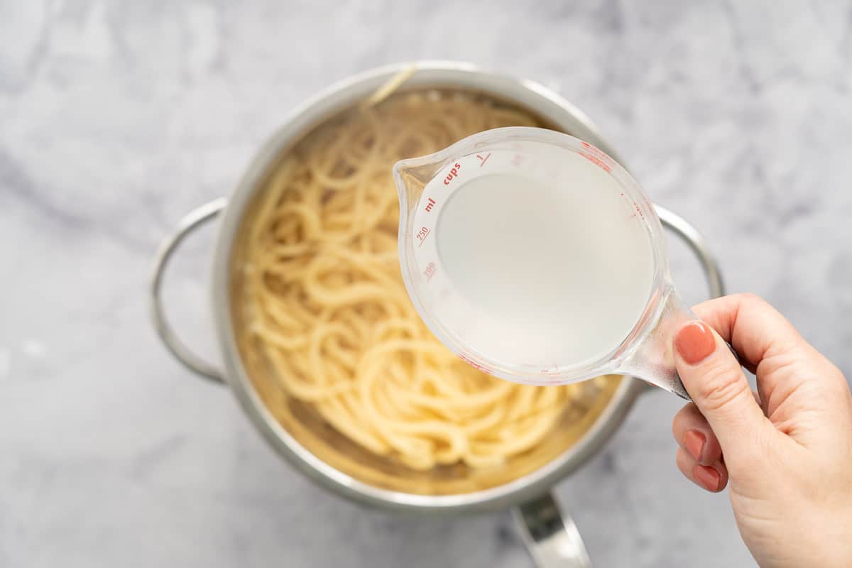 A hand holding a small jug of pasta water above a pot full of cooked spaghetti 