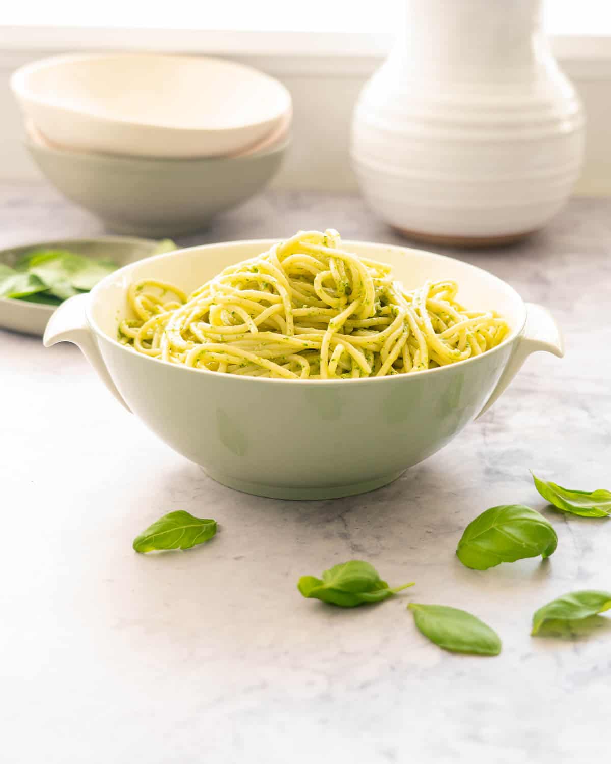 A large bowl filled with pesto pasta sitting on the bench with stacked bowls and a scattering of basil leaves around on the bench 