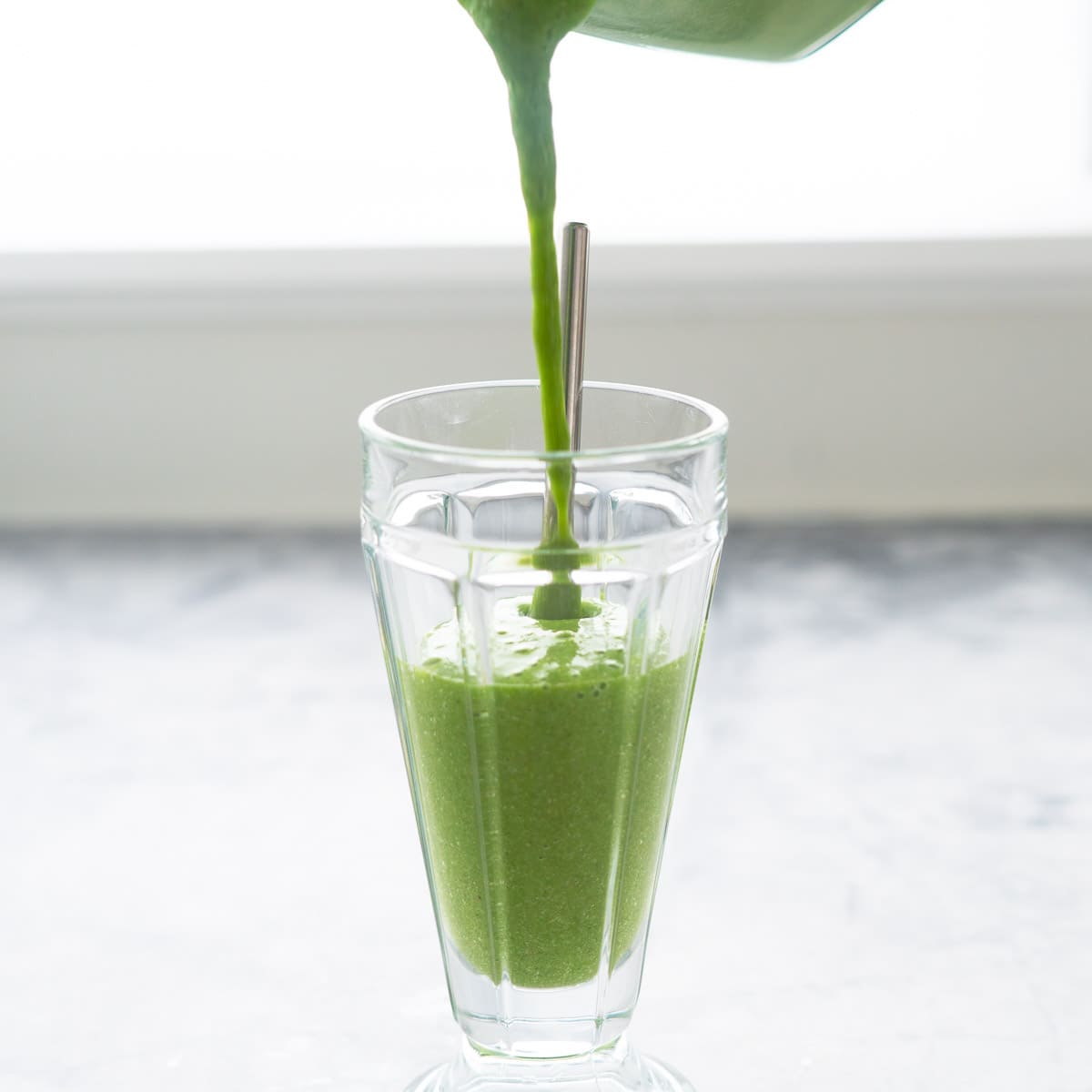 Green spinach smoothie being poured from the blender into a smoothy glass sitting on a bench