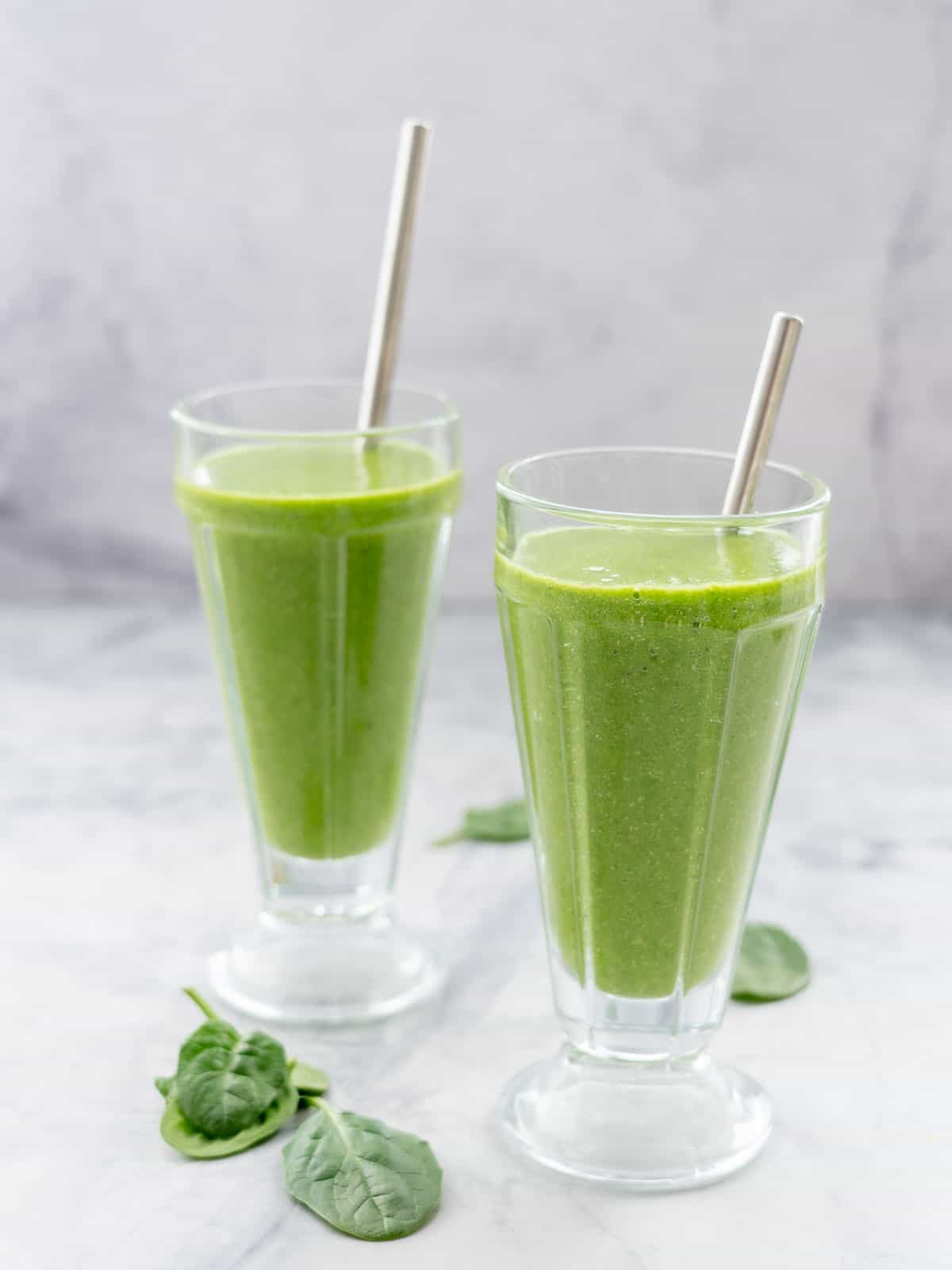 Two tall glasses with metal straws in each of them, filled with bright green smoothie sitting on a bench with a scattering of spinach leaves 