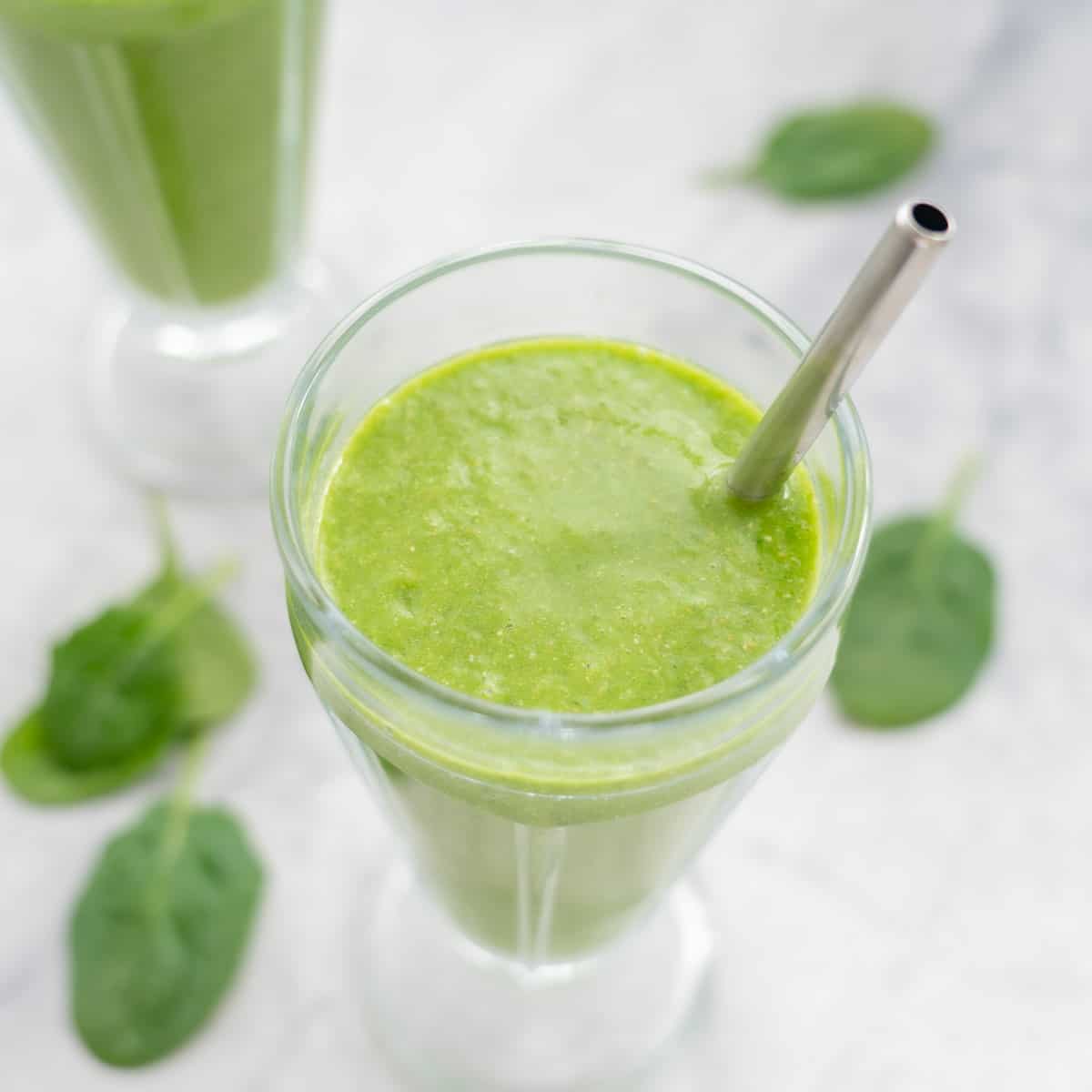 One glass of spinach smoothie placed on the bench with a straw and a scattering of spinach leaves.