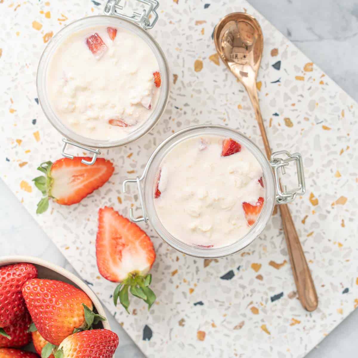 2 jars filled with strawberry overnight oats sitting on a terrazzo serving board next to a brass spoon and sliced strawberry halves and a full bowl of strawberries. 