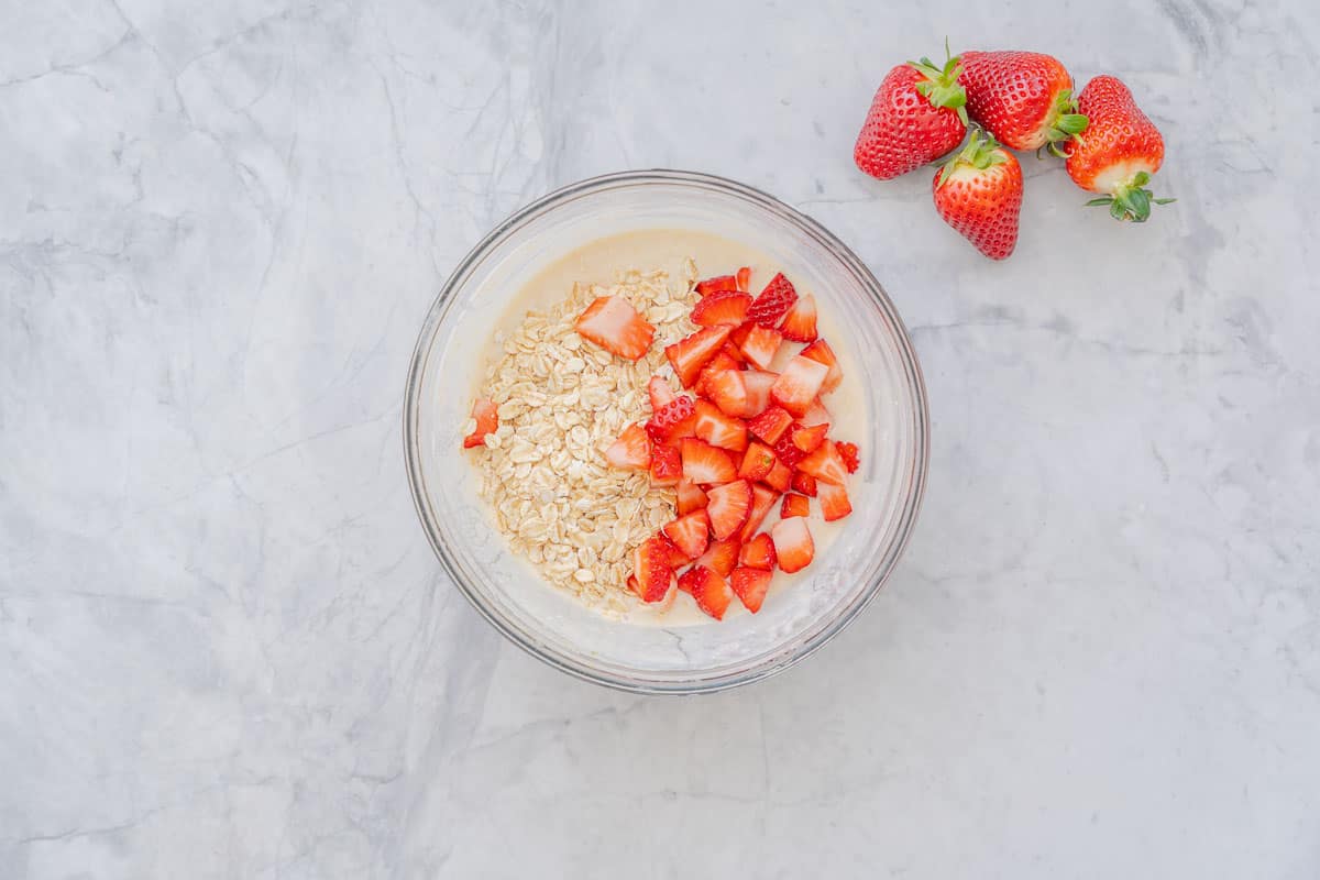 A large glass bowl sitting on the bench filled with ingredients to make the strawberry overnight oats sitting on the bench with a handful of strawberries sitting on the bench to the right 