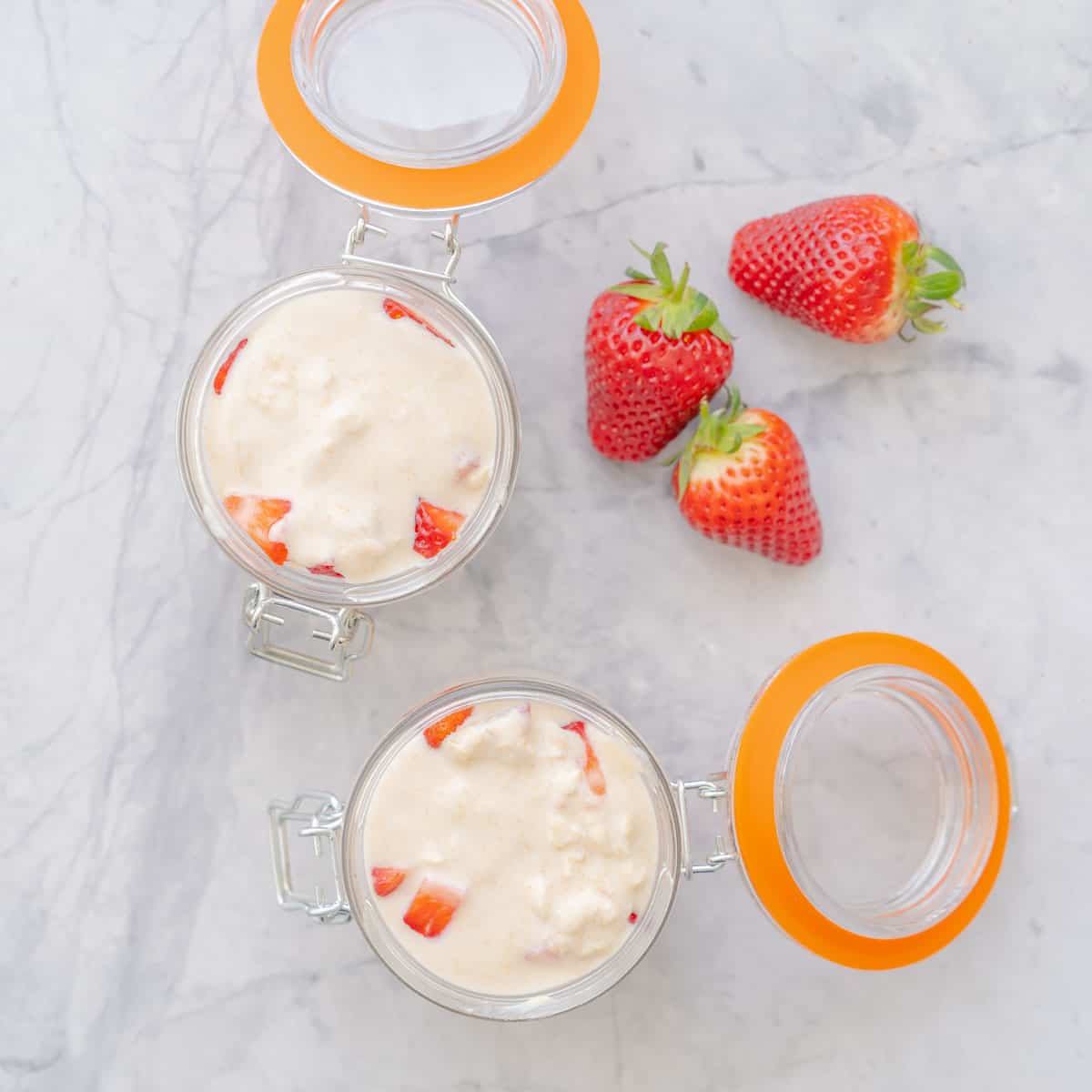 Three strawberries and two small mason jars with their lids open sitting on the bench filled to the brim with overnight oats.