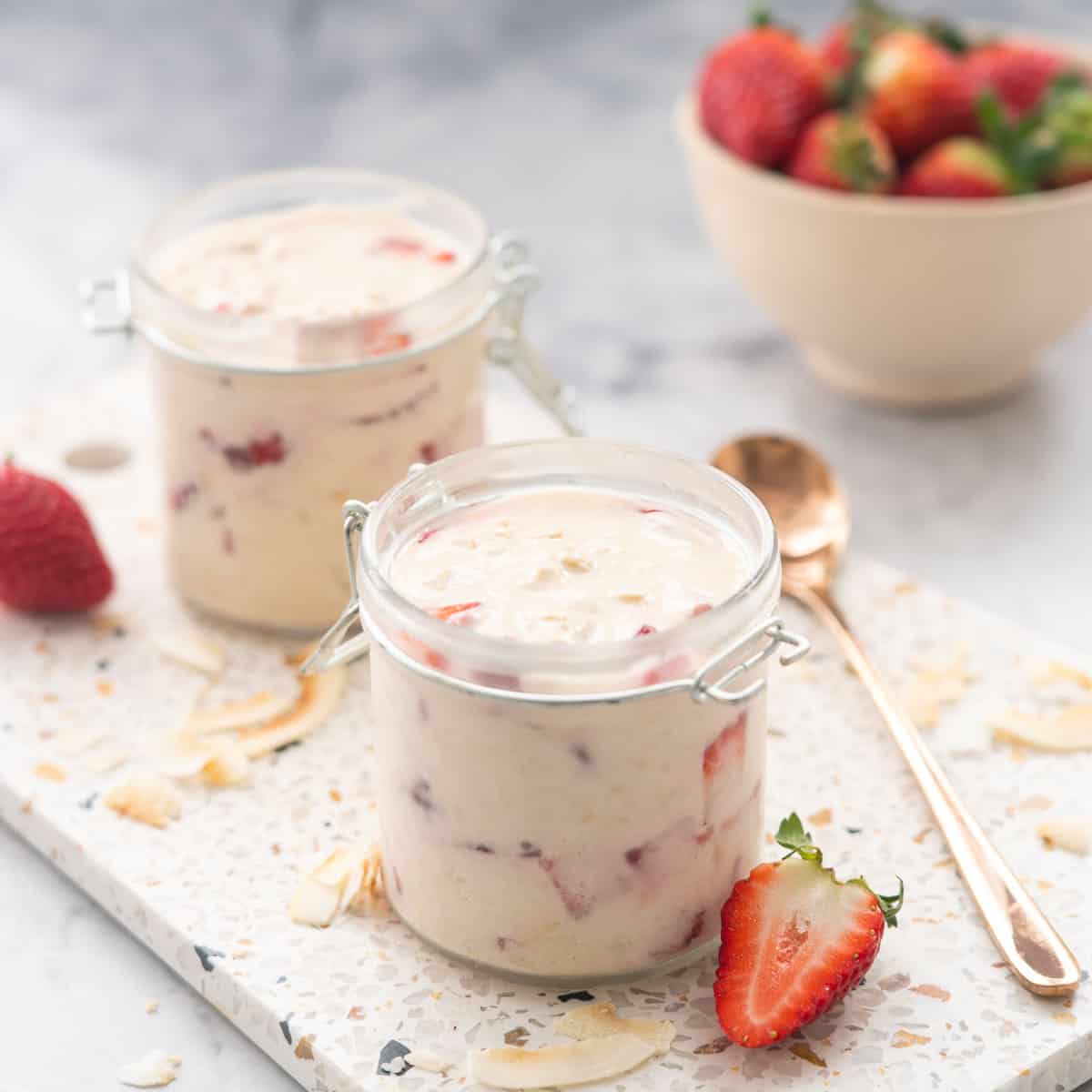 2 jars filled with strawberry overnight oats sitting on a terrazo serving board with a scattering of toasted coconut flakes and strawberry halves 