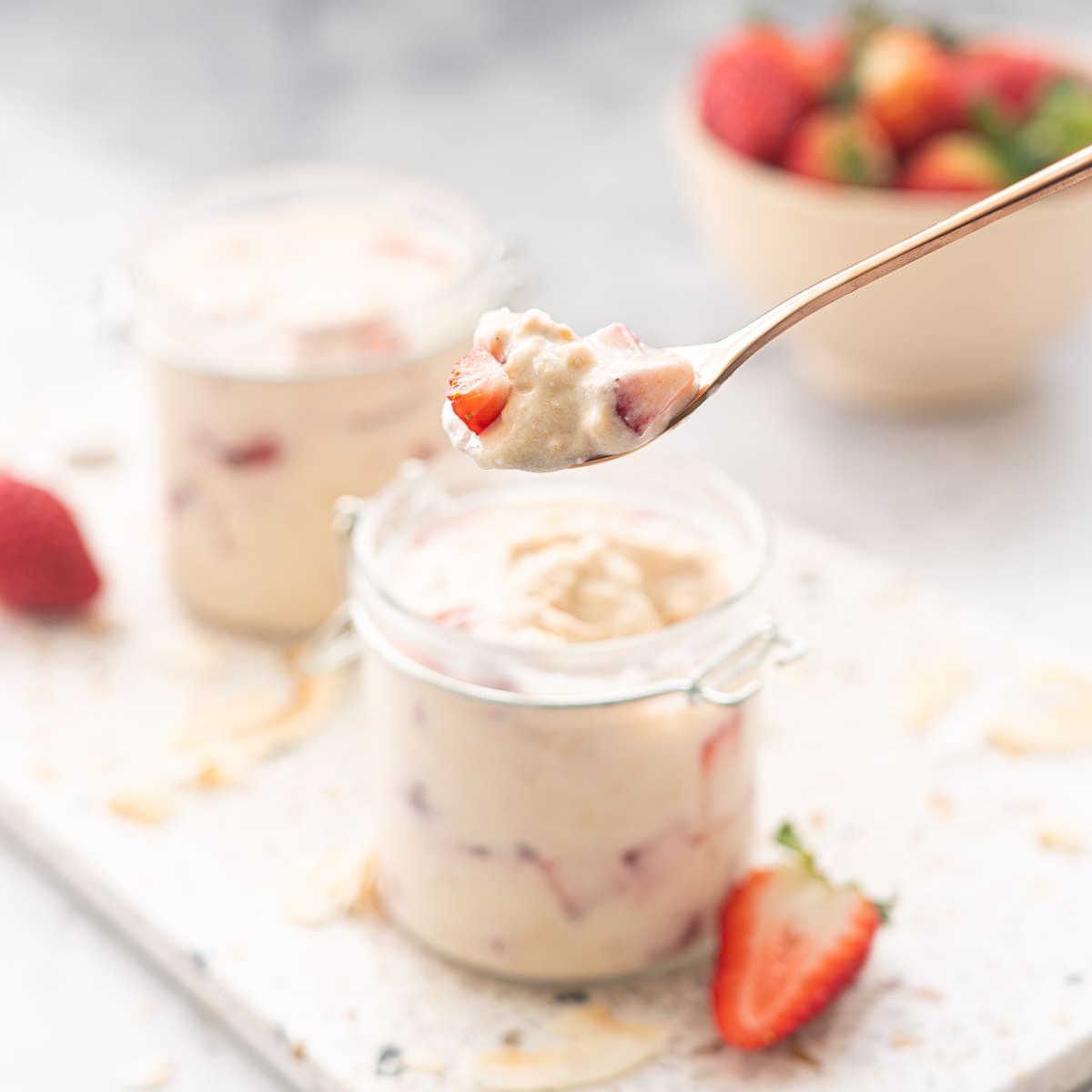 A spoonful of strawberry overnight oats held above 2 full jars  sitting on a terrazzo serving board with a scattering of toasted coconut flakes and strawberry halves  