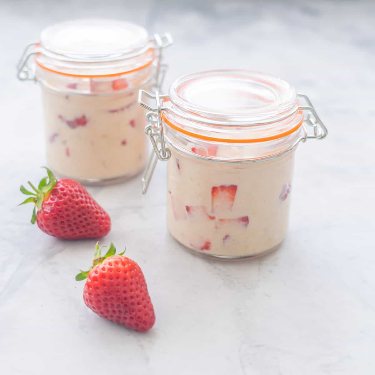 Two mason jars filled with overnight oats sitting on the bench next to two strawberries 