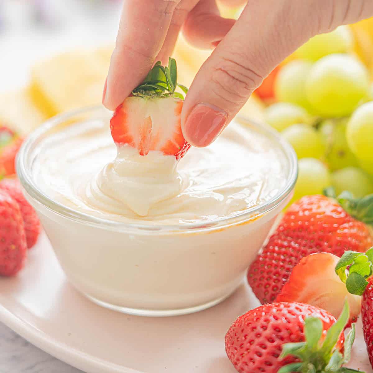 A strawberry being dunked into a glass bowl of Whipped Cottage Cheese surrounded by fresh fruit