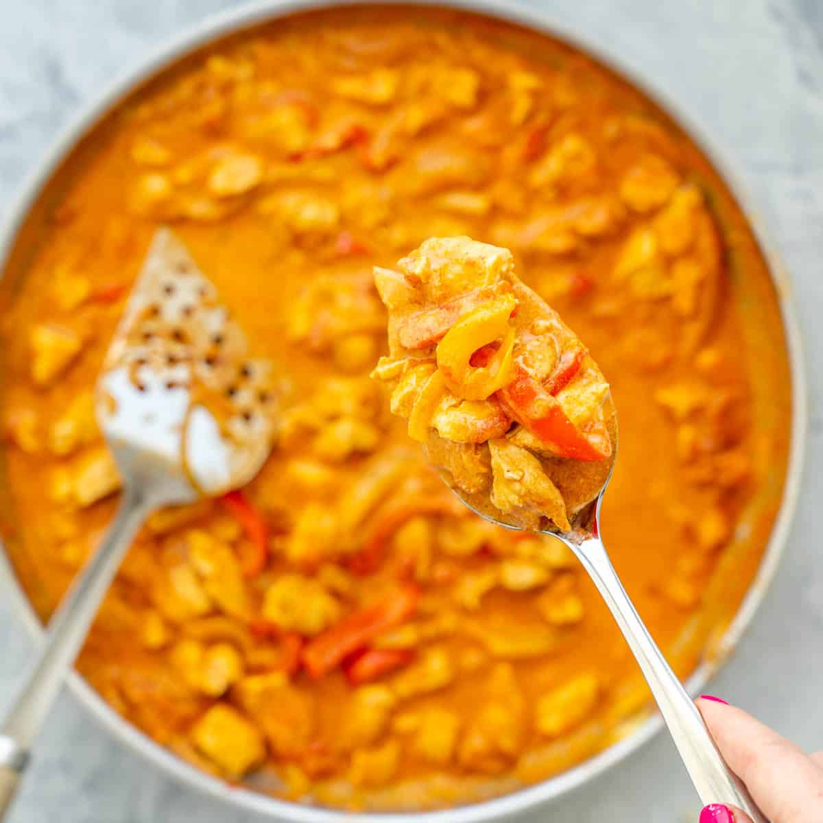 A frying pan full on Chicken Paprikash with a spatula resting on the side and spoonful being held above it. 