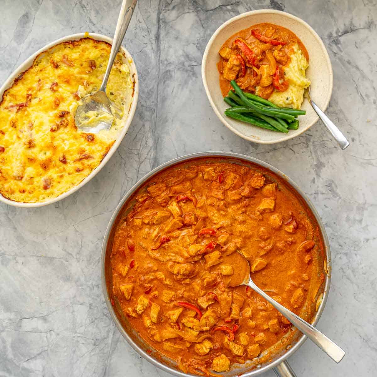 Dishes of Golden brown baked mashed potatoes and chicken Paprikash on the bench next to a serving of the Chicken Parprikash over a big spoonful of the-potatoes. anda side of beans 