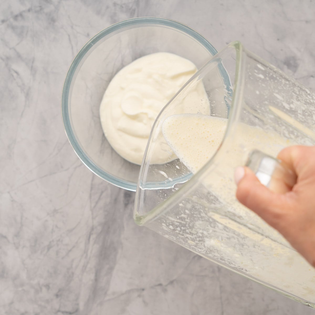 Feta and yoghurt mixture being poured from the blender into a second measure of yoghurt in a glass bowl. 