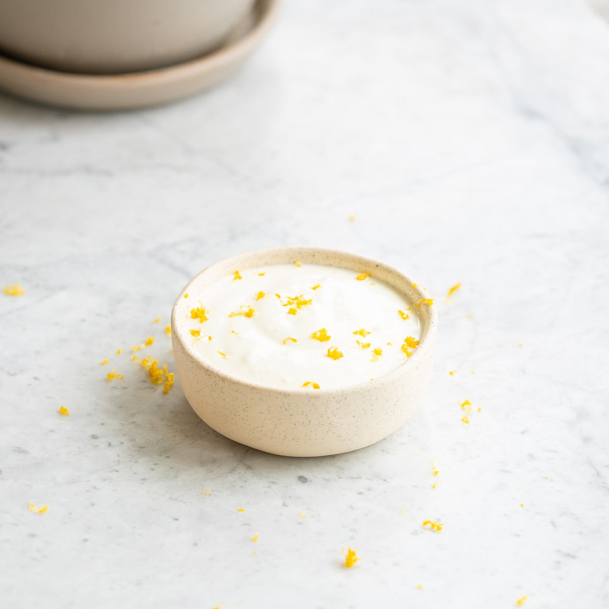 A small ramekin of Lemon Feta Dip with a sprinkling of fresh lemon rind on top sitting on the bench with serving dishes in the background 