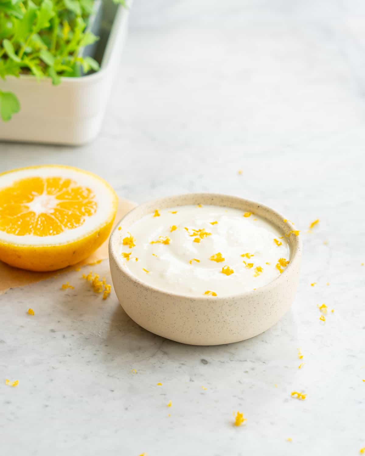 A small ramekin full of lemon feta dip sitting next to a sliced lemon on a bench with a sprinkle of fresh lemon rind 
