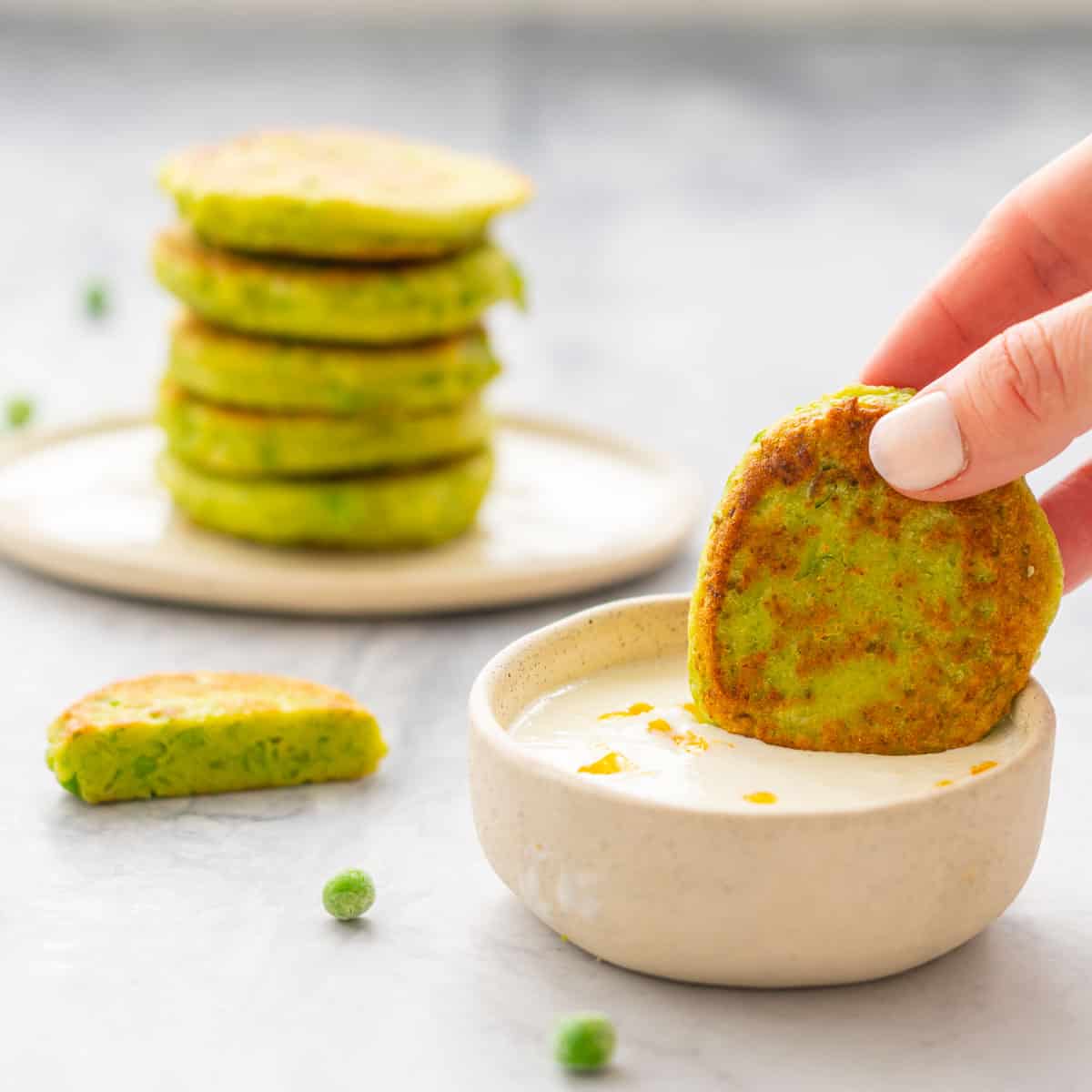 A golden brown pea fritter dunked in the Lemon Feta Dip with a  stack of the fritters in the background on a serving dish and a scatter of peas on the bench  
