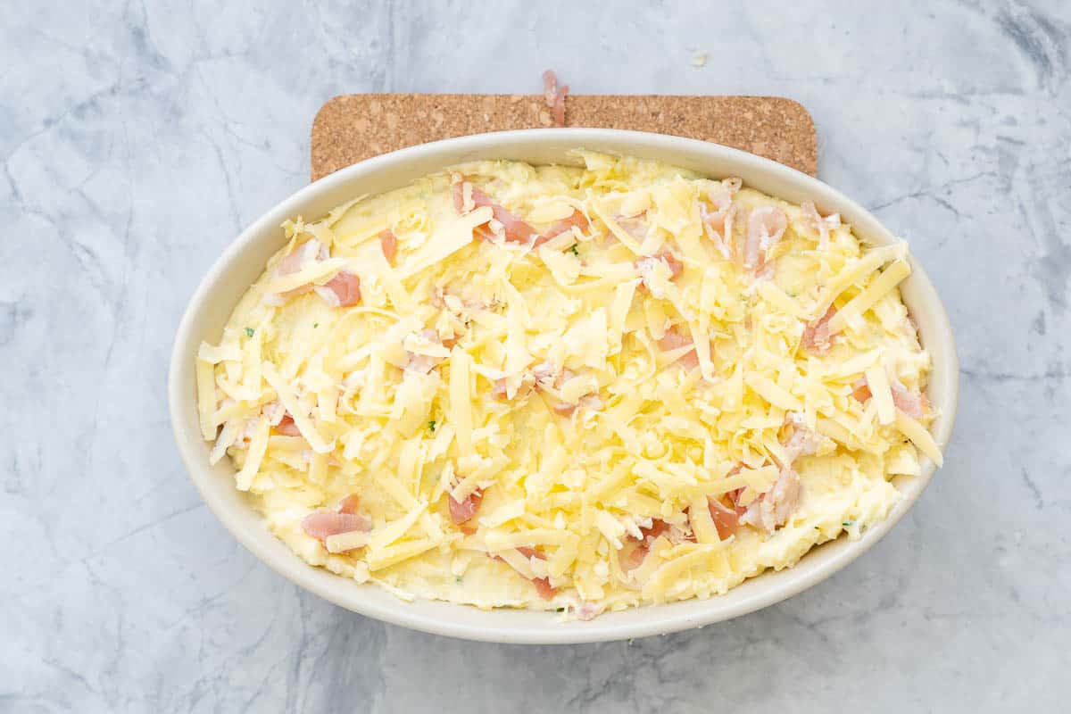 A ceramic dish of mashed potato mixture with bacon and grated cheese on top, sitting on the bench.
