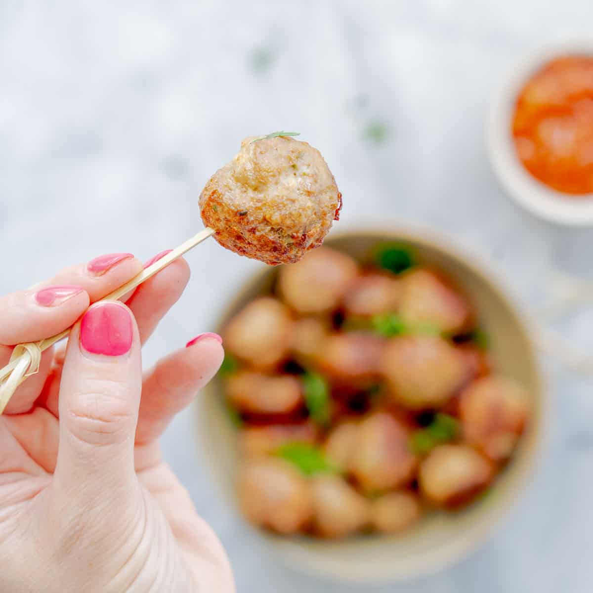 A bowl full of meatballs, with a hand holding up one meatball  above them with a tooth pick.