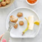 A baked meatball on a baby fork above plate of meatballs and banana.
