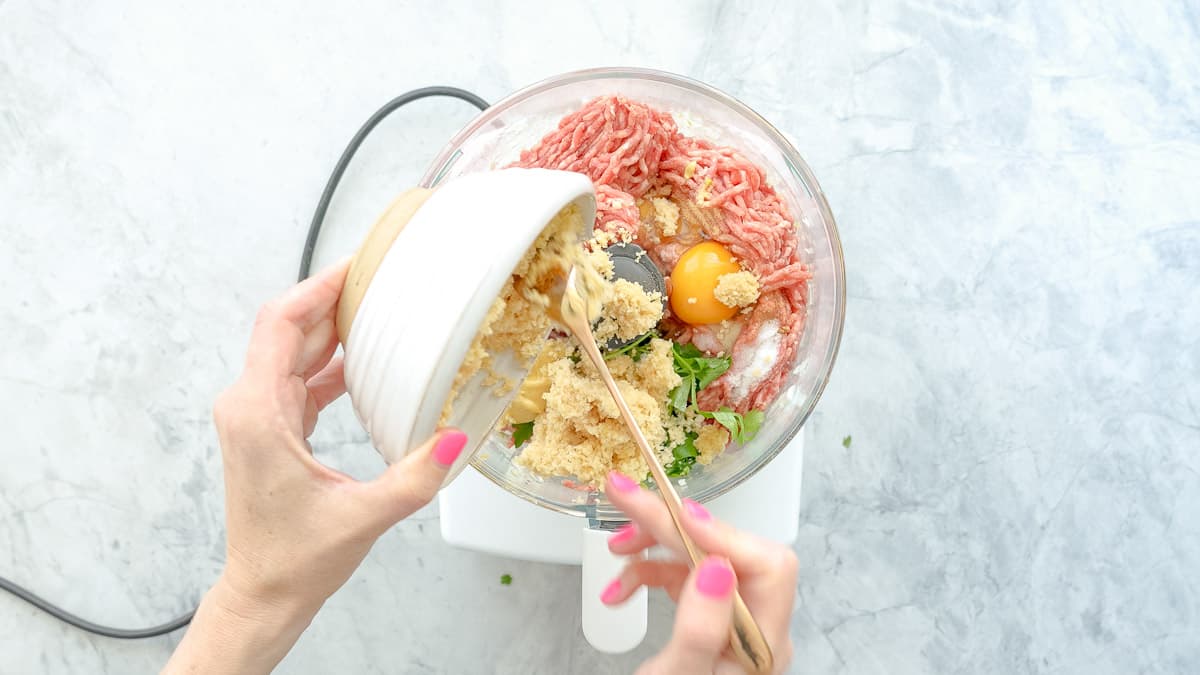All the ingredients placed in a food processor with a hand tipping the panko breadcrumbs into it from a small ceramic bowl