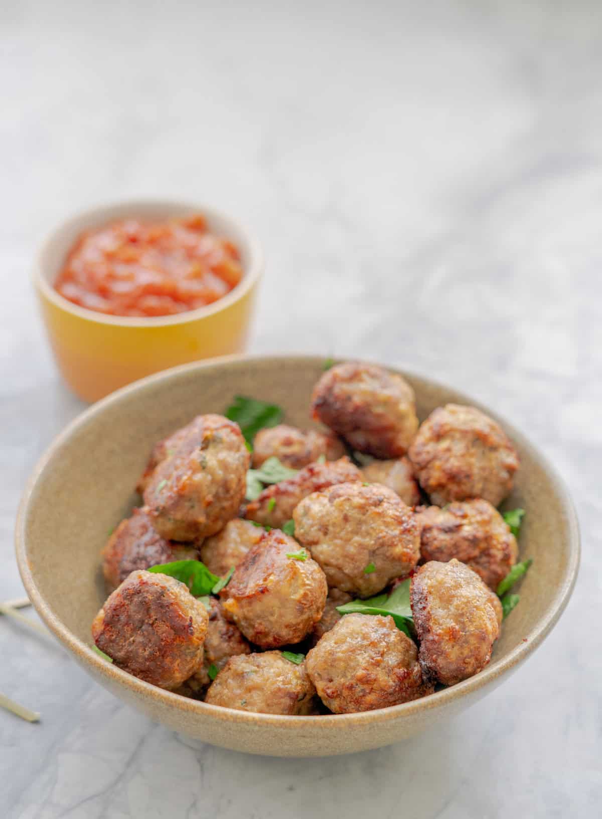 A ceramic serving bowl full of baked meatballs with a sprinkle of parsley next to a small ceramic serving dish of chutney