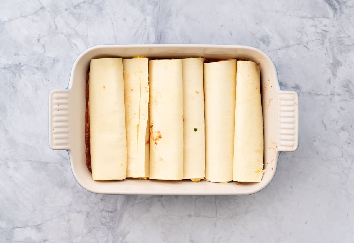 A baking dish sitting on a  bench with 6 stuffed pasta rolls laid out in the bottom of it. 