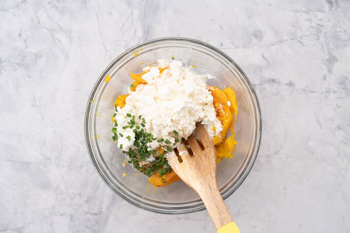 Roasted butternut,  cottage cheese, chives and salt and pepper in a glass mixing bowl with a wooden spoon resting on the side.