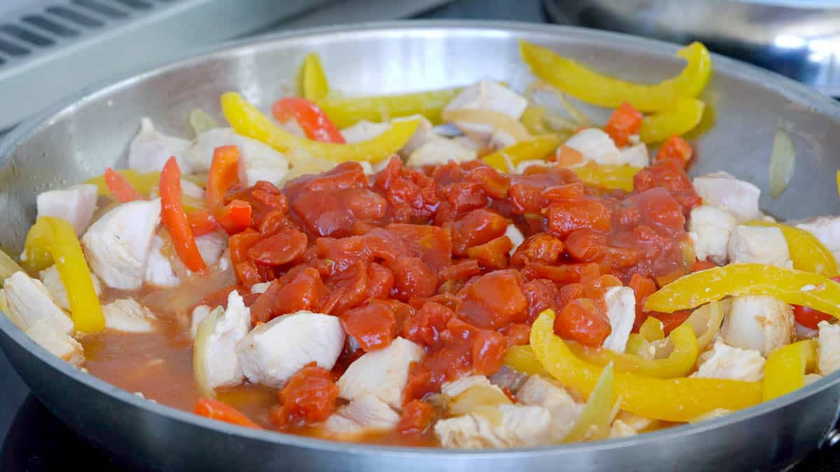 Sliced capsicum onions and chicken sautéing in an oiled frying pan with a tin of tomatoes added.