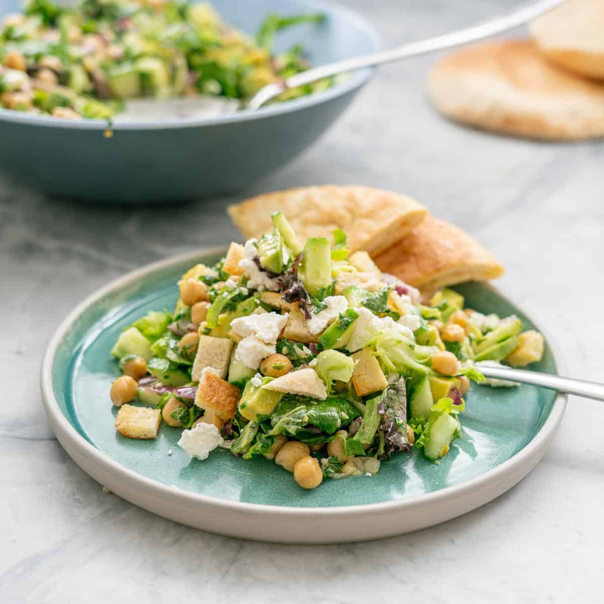 A salad of chickpeas, avocado, feta and green vegetables on a turquoise plate with wedges of pita bread. 
