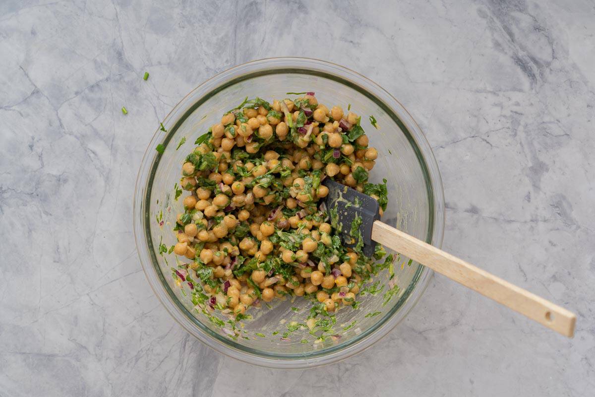 A large glass mixing bowl of chickpeas, chopped red onion, chopped parsley and salad dressing. 