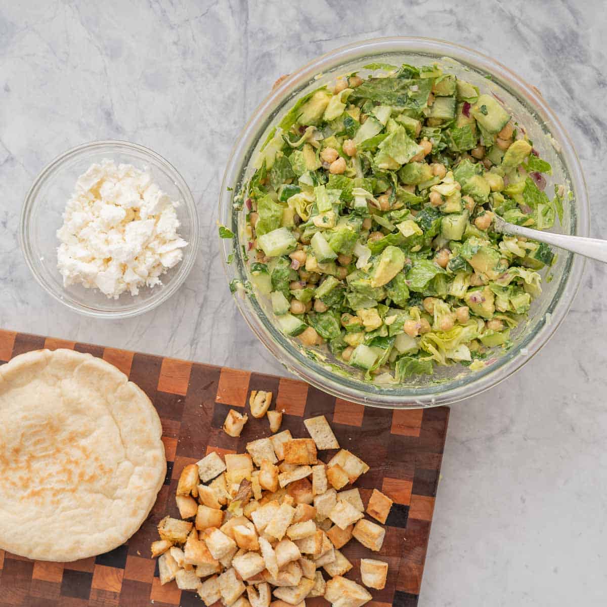 A large glass bowl of chickpea avocado salad on a bench  with a small bowl crumbled feta and pita bread. 
