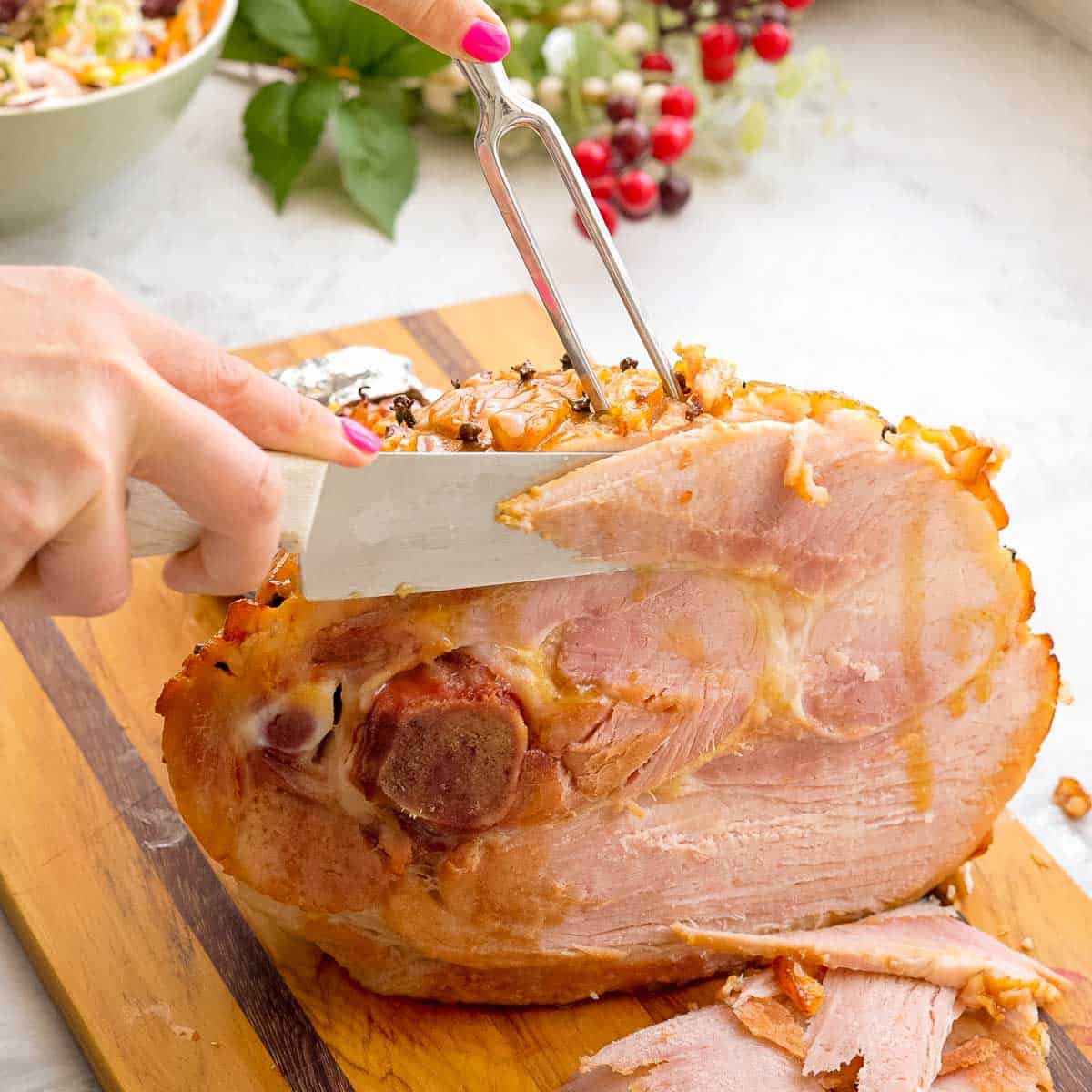 A large cooked ham being sliced by a large knife on a wooden chopping board with multiple slices beside it. 