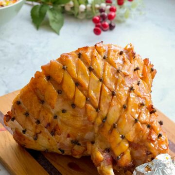 A cooked glazed ham with cloves pressed into the corners of diamond shaped cuts in the fat layer resting on a wooden chopping board on the bench with christmasy decorations in the background