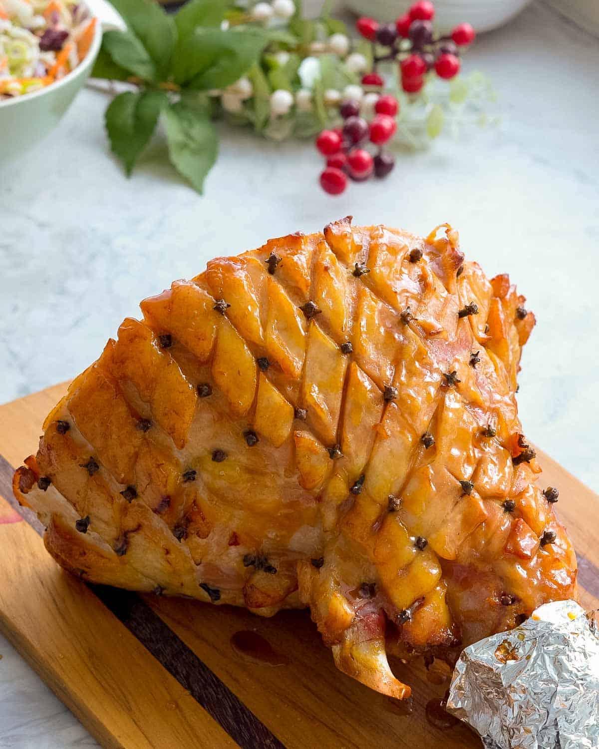 A shoulder of glazed ham, with a piece of tinfoil wrapped around the end sitting on a wooden chopping board