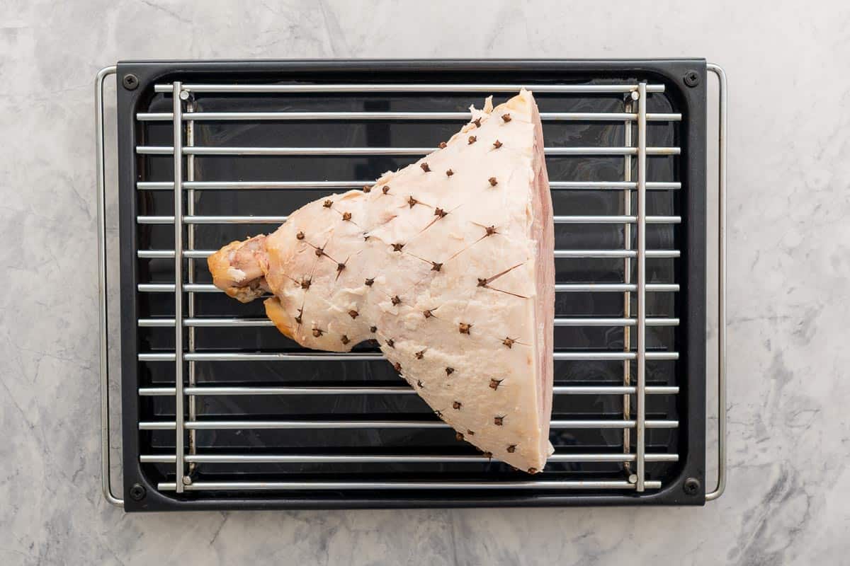 Diamond-shaped cuts in the fat layer of a large uncooked ham with cloves pressed between each corner which is sitting on a rack in a big oven dish