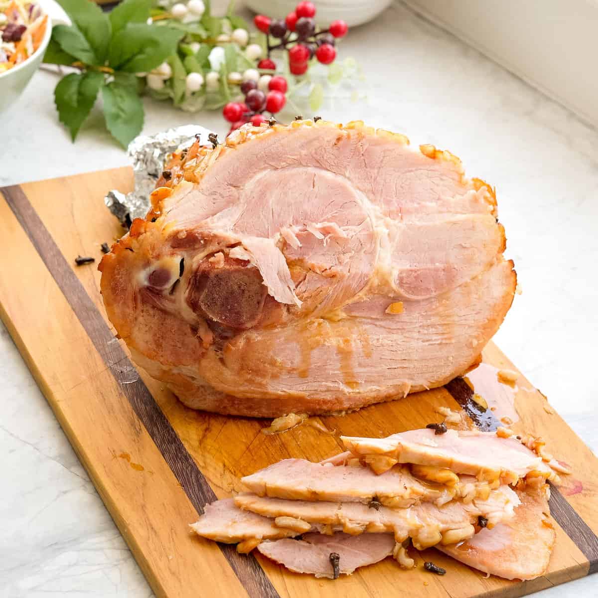 A large cooked ham resting on a wooden chopping board  on the bench with multiple slices of ham beside it. 