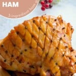 A cooked glazed ham with cloves pressed into the corners of diamond shaped cuts in the fat layer resting on a wooden chopping board on the bench with christmasy decorations in the background with text overlay for pinterest