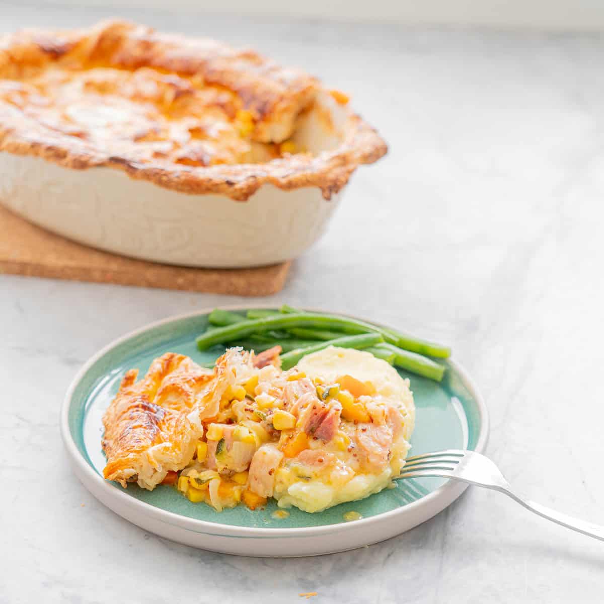Golden brown baked ham pot pie resting on a chopping board, behind a serving with a side of beans on a teal coloured ceramic plate and a fork resting on the side