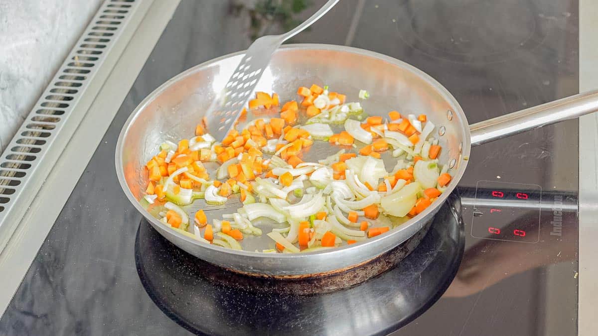 A frying pan sautéing onions and carrots on an element with a spatula stirring it.