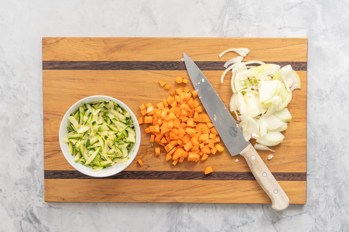 A wooden chopping board and knife on the bench with diced carrots, sliced onions and a white bowl full of grated zucchini 